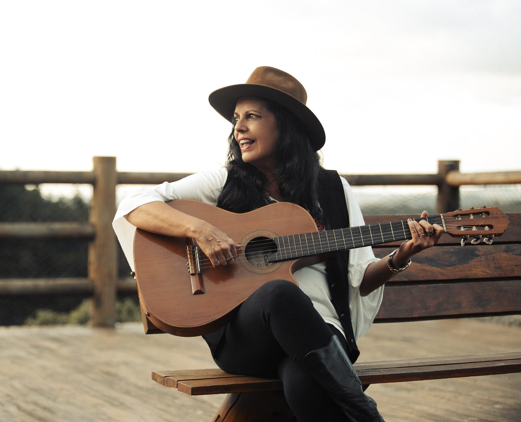 Waleska Santos sentada num banco de madeira, de chapéu, cantando e tocando violão