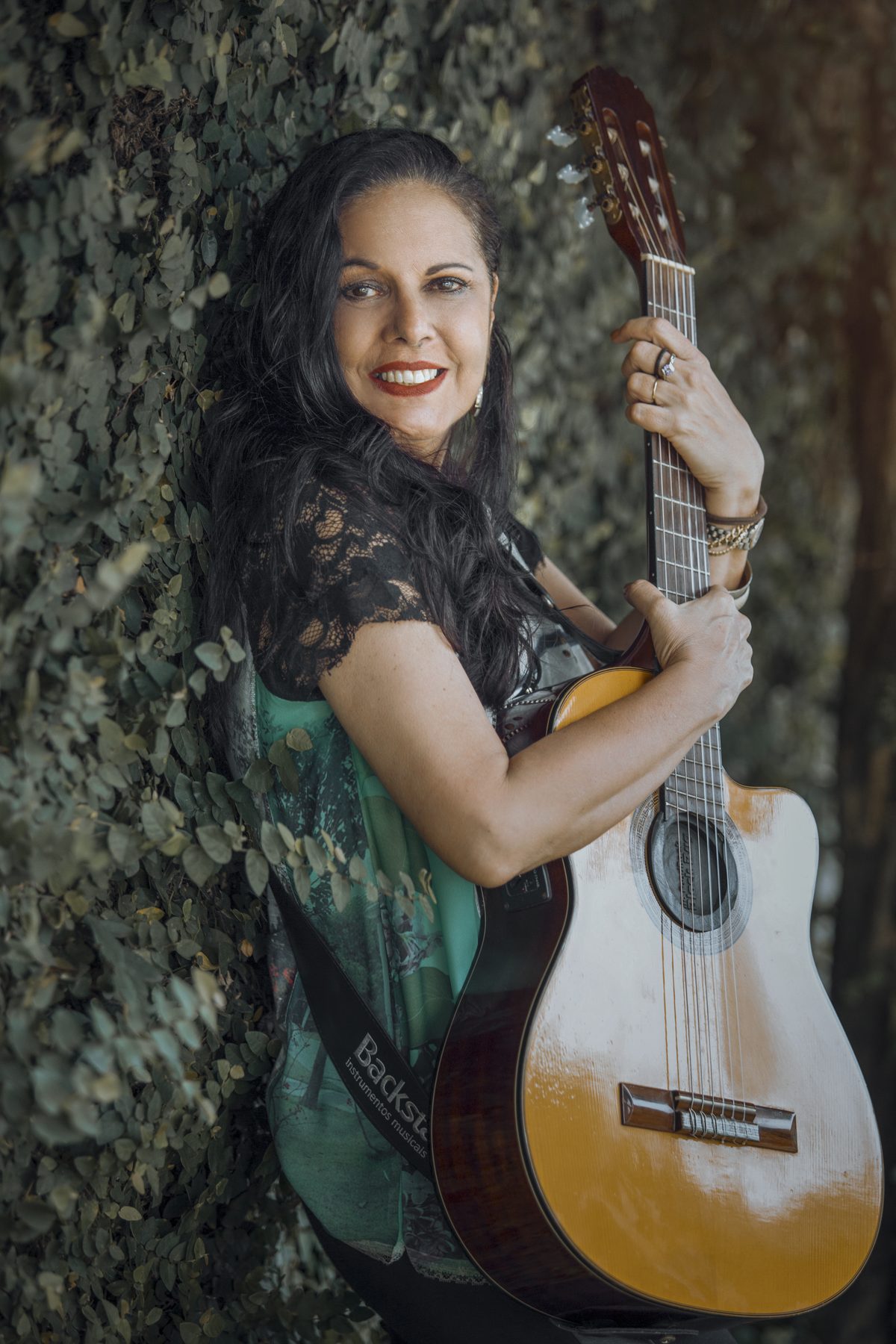 Waleska sorrindo, encostada num muro de folhas, com o violão no colo